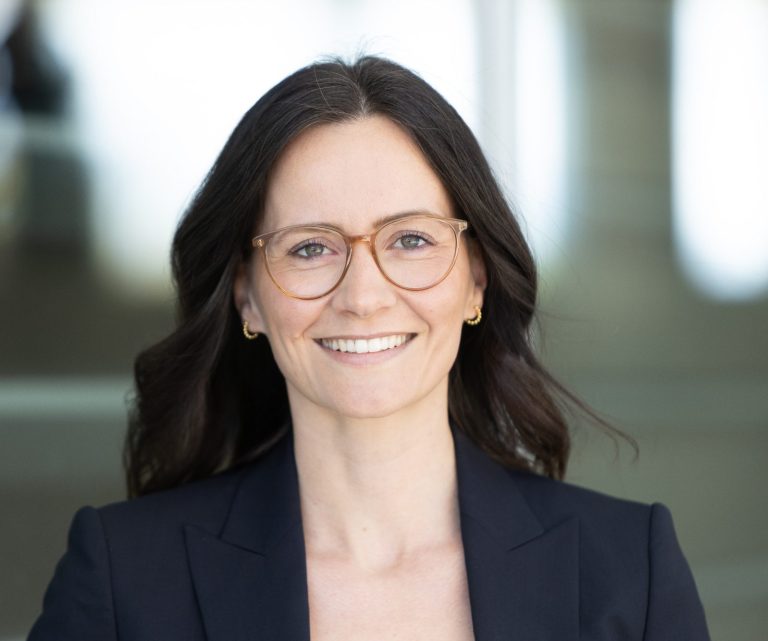 Sarah Wütz Damenportrait mit langen, dunklen Haaren und Brille, in Büroatmosphäre.